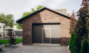 Nowoczesny, ceglany garaż do zgłoszenia, z szarą bramą i drzwiami. Przed nim podjazd z kostki brukowej, po bokach drzewa i plac zabaw.