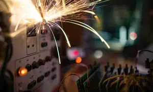 Iskry lecą z panelu sterowania, sugerując **przeciążenie instalacji elektrycznej**. W tle widać płytki drukowane i rozmyte światła.