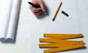Rysowanie planów domu, ołówek, linijka i zwinięte projekty. Zastanawiasz się, ile trwa budowa domu? Czas zależy od wielu czynników.