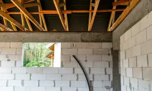 Budowa domu w toku. Widoczny szalunek wieńca nad ścianami z bloczków, drewniana konstrukcja dachu i otwór okienny z widokiem na las.
