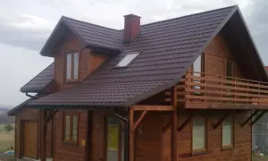 Drewniany dom szkieletowy z balkonem i brązowym dachem, otoczony zielenią.