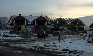 Budowa domów na gruntach ornych. Widać zarys budynków, dachy, rusztowania i samochód.