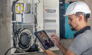Technik w kasku i okularach ochronnych sprawdza przegląd instalacji elektrycznej domu na tablecie. Widoczne przewody i rozdzielnica.