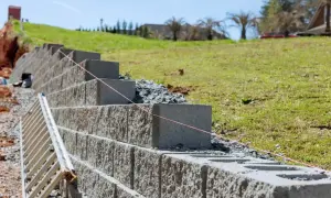 Budowa muru oporowego w ogrodzie. Betonowe bloki układane są warstwami, tworząc solidną konstrukcję. W tle zielona skarpa i dom.