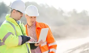 Dwóch mężczyzn w kaskach i kamizelkach odblaskowych analizuje projekt na laptopie. Jeden z nich, kierownik budowy, wskazuje coś na ekranie.