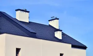 Nowoczesny dach bezokapowy z ciemnogranatową dachówką i białymi kominami, pod błękitnym niebem.