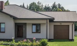 Dach domu pokryty ciemną blachą trapezową, która stanowi jeden z rodzajów blachy trapezowej. Widać też komin i okna.