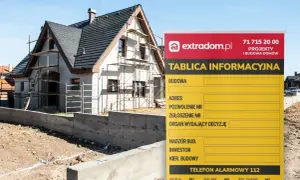 Tablica informacyjna budowy z danymi kontaktowymi i numerami alarmowymi, obok dom w trakcie budowy. RODO na budowie.