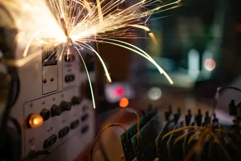 Iskry lecą z panelu sterowania, sugerując **przeciążenie instalacji elektrycznej**. W tle widać płytki drukowane i rozmyte światła.