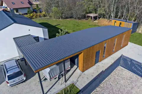 Nowoczesny budynek z drewnianą elewacją i ciemnym, **dachem jednospadowym**. Obok stoi samochód i przyczepa pod zadaszeniem.