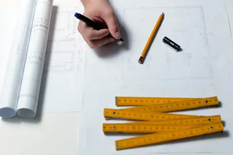 Rysowanie planów domu, ołówek, linijka i zwinięte projekty. Zastanawiasz się, ile trwa budowa domu? Czas zależy od wielu czynników.