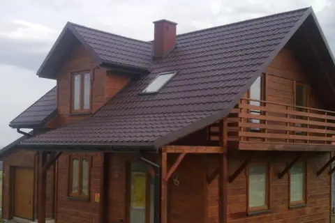Drewniany dom szkieletowy z balkonem i brązowym dachem, otoczony zielenią.