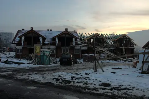 Budowa domów na gruntach ornych. Widać zarys budynków, dachy, rusztowania i samochód.