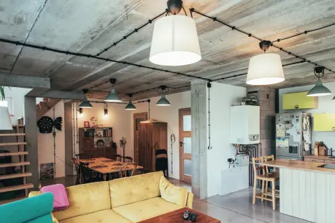 Nowoczesne wnętrze z betonowym sufitem i widoczną instalacją natynkową w rurkach. Wygodna kanapa, jadalnia i kuchnia tworzą funkcjonalną przestrzeń.