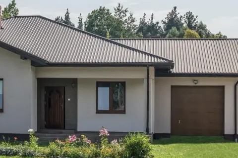 Dach domu pokryty ciemną blachą trapezową, która stanowi jeden z rodzajów blachy trapezowej. Widać też komin i okna.