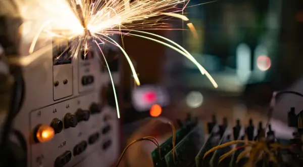 Iskry lecą z panelu sterowania, sugerując **przeciążenie instalacji elektrycznej**. W tle widać płytki drukowane i rozmyte światła.