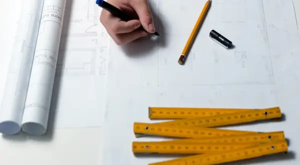 Rysowanie planów domu, ołówek, linijka i zwinięte projekty. Zastanawiasz się, ile trwa budowa domu? Czas zależy od wielu czynników.