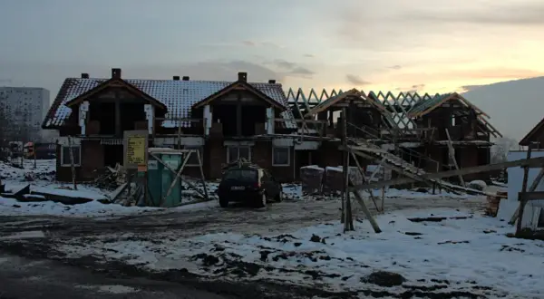 Budowa domów na gruntach ornych. Widać zarys budynków, dachy, rusztowania i samochód.