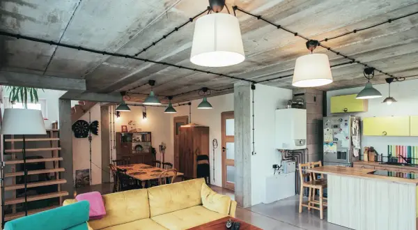 Nowoczesne wnętrze z betonowym sufitem i widoczną instalacją natynkową w rurkach. Wygodna kanapa, jadalnia i kuchnia tworzą funkcjonalną przestrzeń.