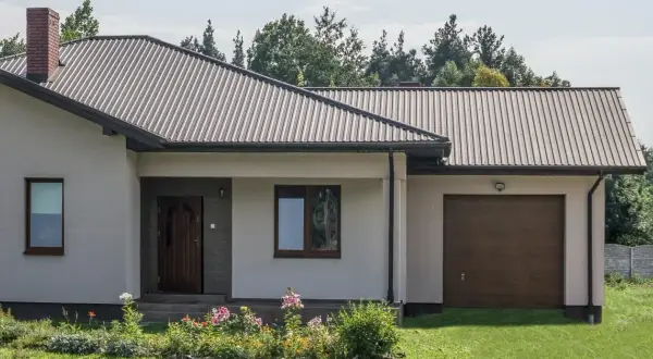 Dach domu pokryty ciemną blachą trapezową, która stanowi jeden z rodzajów blachy trapezowej. Widać też komin i okna.