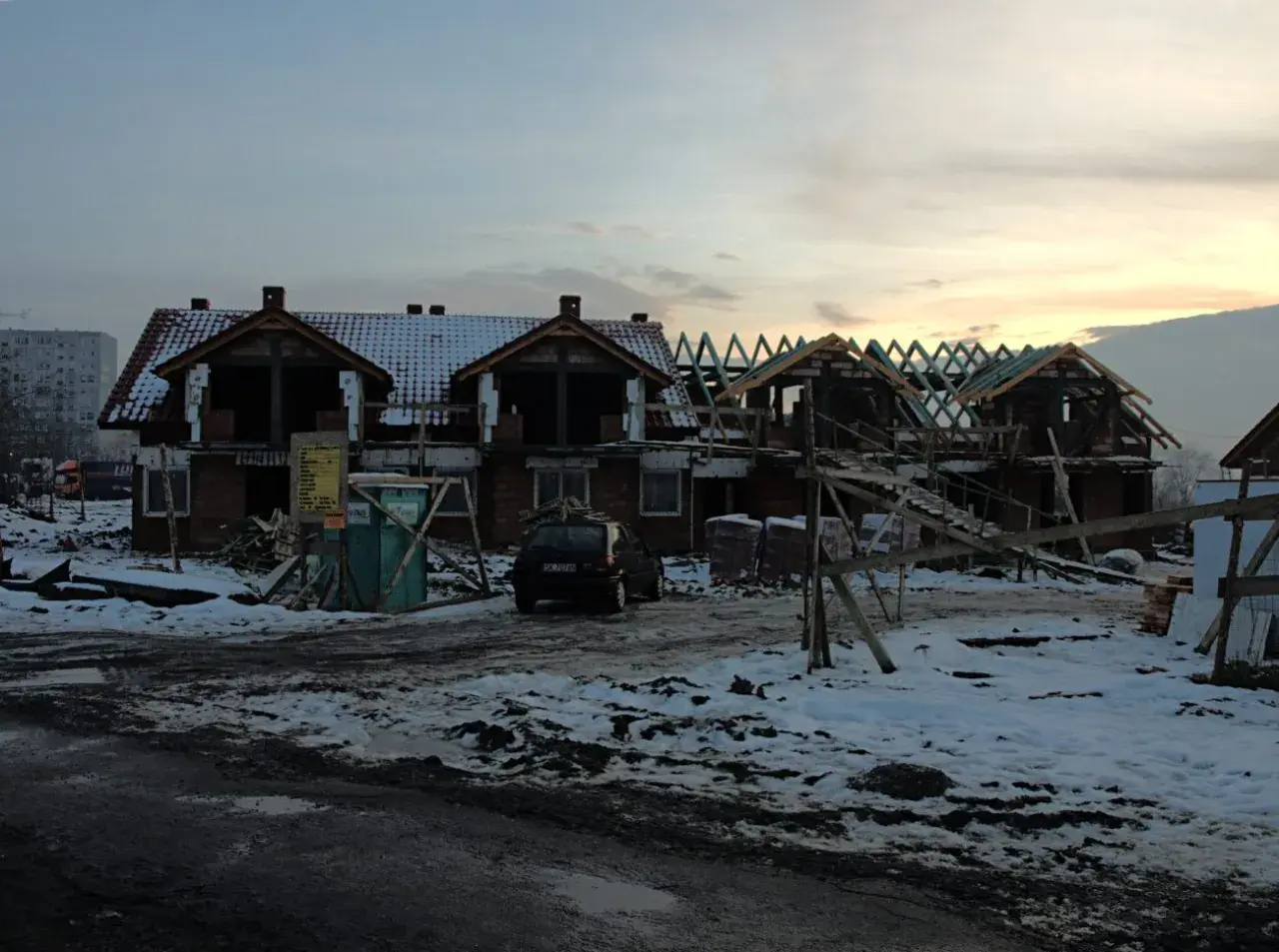 Budowa domów na gruntach ornych. Widać zarys budynków, dachy, rusztowania i samochód.