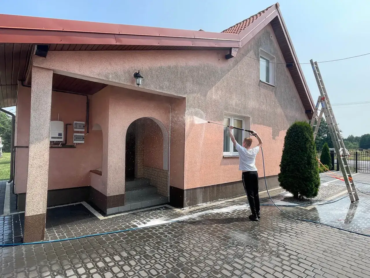 Chłopak myje elewację domu myjką ciśnieniową, tworząc pionowe pasy na ścianie.