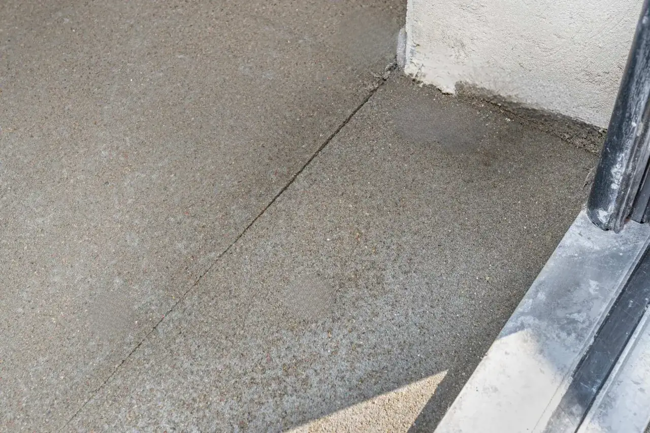 Szara, chropowata powierzchnia betonu z widoczną szczeliną, kt&oacute;ra może być jednym z rodzaj&oacute;w dylatacji. Obok znajduje się fragment ściany i metalowa poręcz.