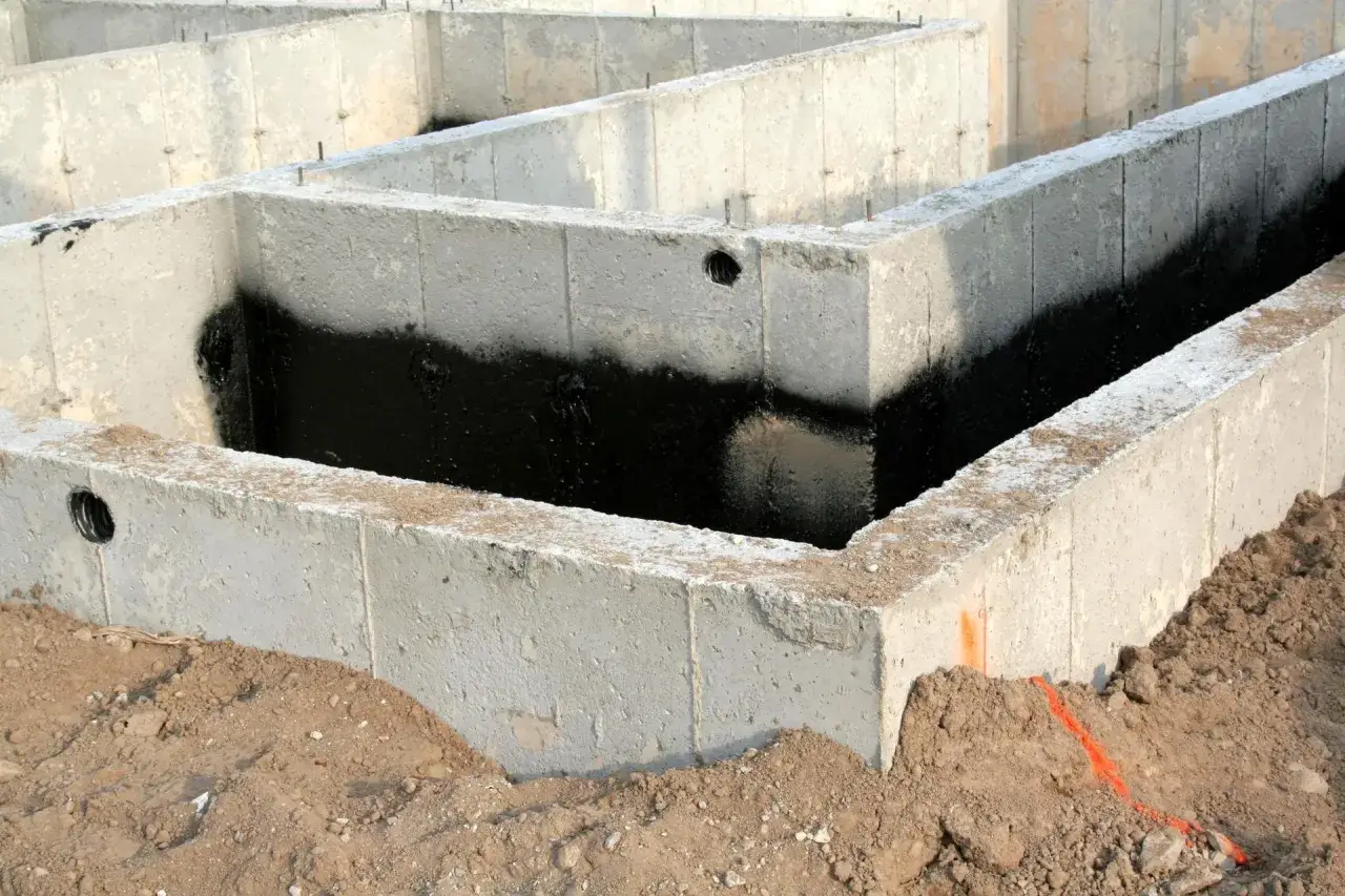 Fundamenty domu w budowie. Widoczny mur z bloczk&oacute;w betonowych, częściowo pokryty czarną izolacją przeciwwilgociową.