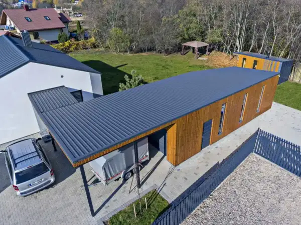 Nowoczesny budynek z drewnianą elewacją i ciemnym, **dachem jednospadowym**. Obok stoi samochód i przyczepa pod zadaszeniem.
