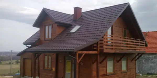 Drewniany dom szkieletowy z balkonem i brązowym dachem, otoczony zielenią.