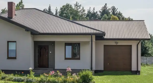 Dach domu pokryty ciemną blachą trapezową, która stanowi jeden z rodzajów blachy trapezowej. Widać też komin i okna.